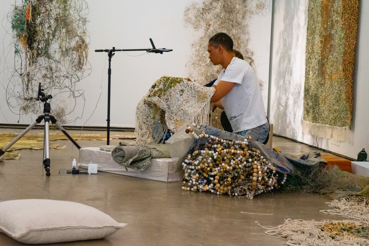 Event photograph from ‘Knots’, a conversation between Igshaan Adams and Josh Ginsburg, marking the close of ‘Open Production’, Adams’ hybrid studio/exhibition in A4’s Gallery. On the right, Adams and Ginsburg examine one of Adams’ woven tapestries under a recording device mounted on a stand.
