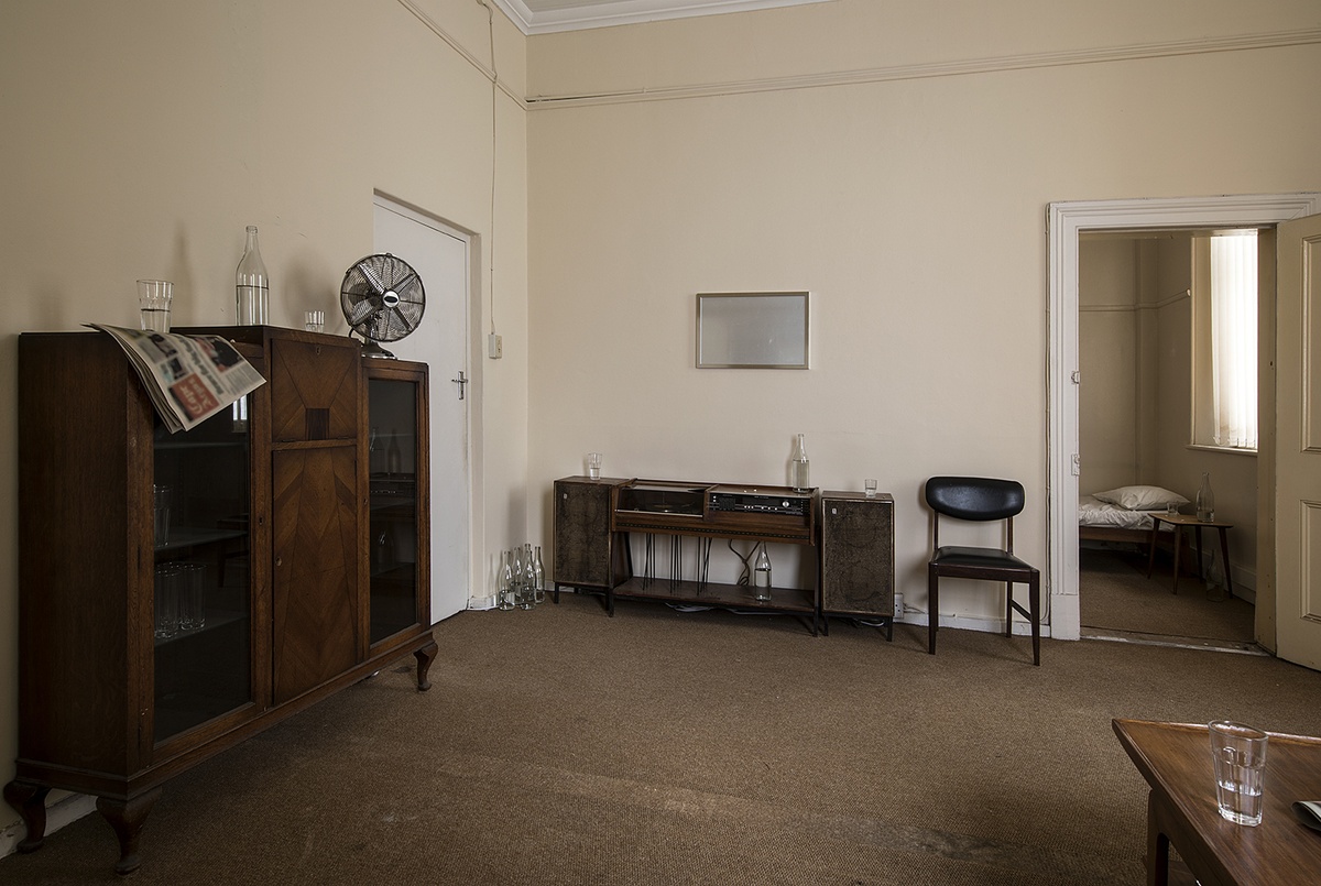 Installation photograph from Gain Maria Tosatti’s offsite exhibition ‘My Hart is so Leeg soon ’n Spieël (My Heart is a Void, the Void is a Mirror)’. On the left, a wooden display case with a glass bottle, drinking glasses, printed matter and electronic fan on top of it. At the back, a wooden record player console with glass bottles and drinking glasses, and a black chair on the right.
