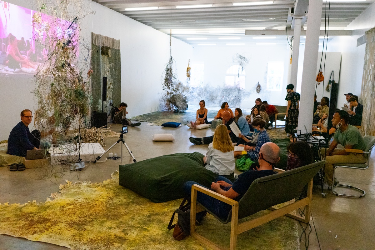 Event photograph from ‘Knots’, a conversation between Igshaan Adams and Josh Ginsburg, marking the close of ‘Open Production’, Adams’ hybrid studio/exhibition in A4’s Gallery. On the left, Ginsburg and Adams are seated among Adams’ works-in-progress, with a live video projection on the wall behind them. On the right, attendees are seated on cushions, chairs and couches.
