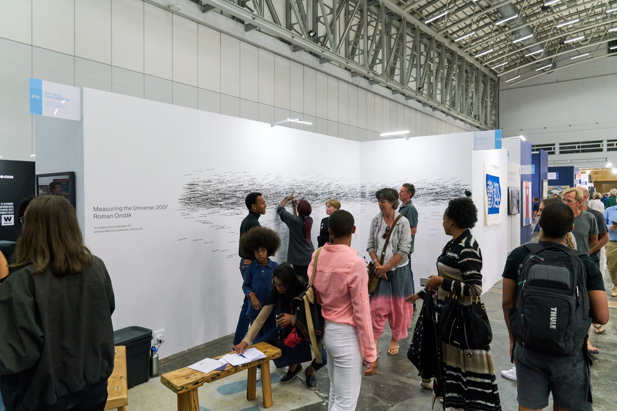 Event photograph of Roman Ondák’s performance piece ‘Measuring the Universe’ at A4’s booth at the 2020 Cape Town Art Fair. At the back, a white wall is covered with overlapping marks that indicate participant’s height, names and the date of their participation in black felt pen. At the front, A4’s Obakeng Motsepe and group of fair attendees.
