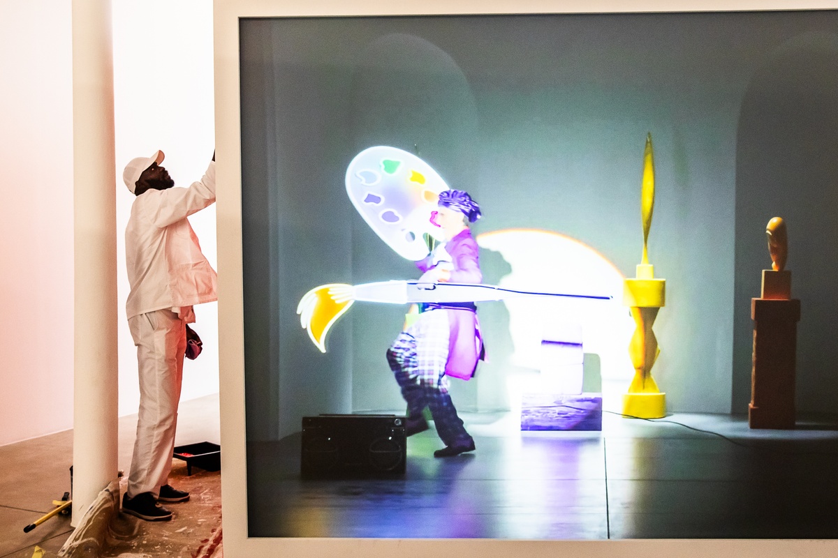 Event photograph from the preview of the 'A Little After This' exhibition in A4 Arts Foundation. On the right, a large white wooden box with a back-projected screen displays the video from Alex Da Corte's 'ROY G BIV' installation. On the left, an individual dressed in white overalls repaints the box red.
