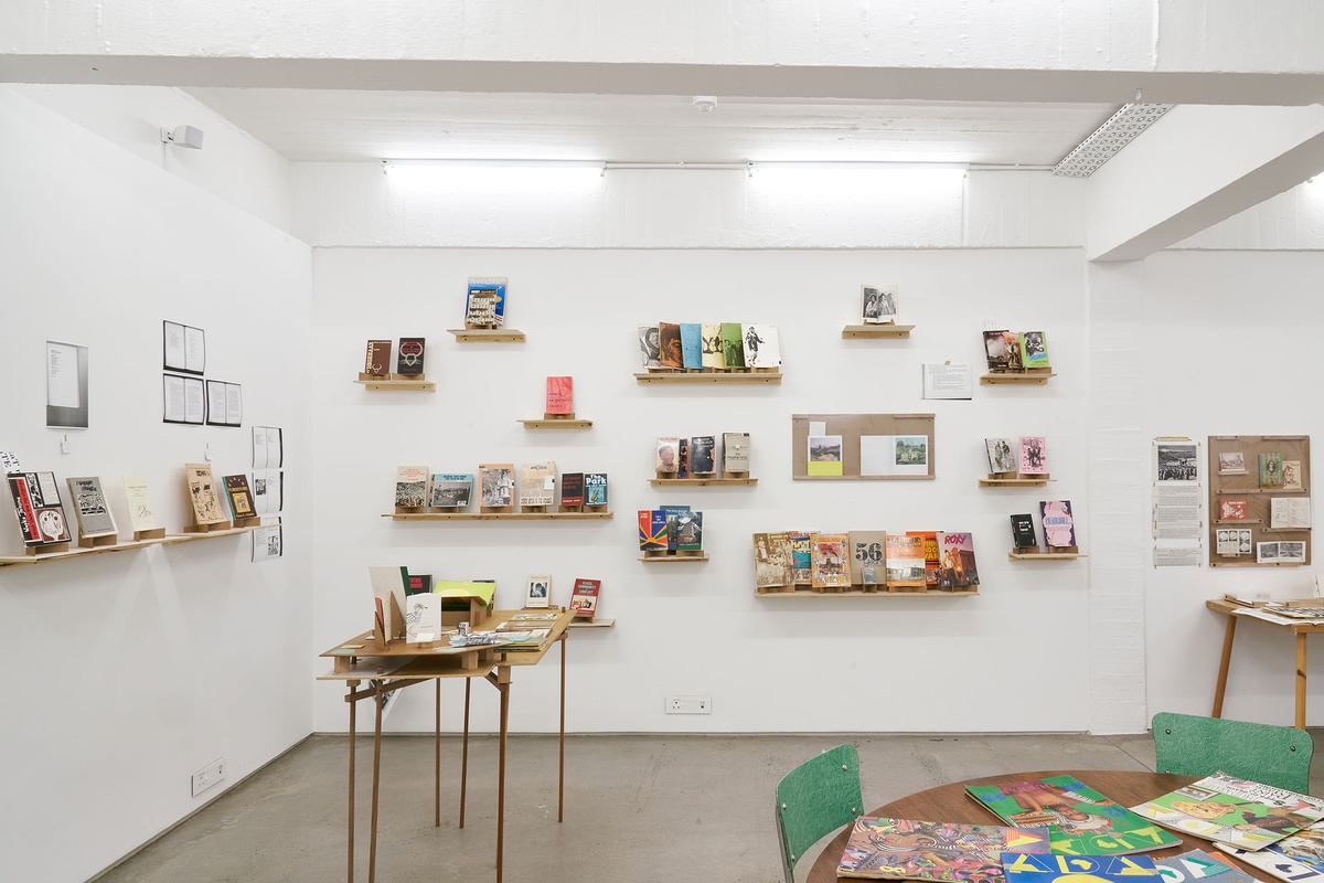 Installation photograph from the Papertrails exhibition in A4’s Reading Room. On the left, a wooden table with printed matter. At the back, various wall-mounted shelves with printed matter.
