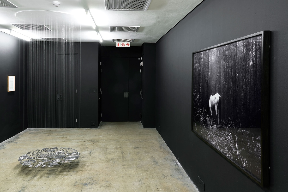 Installation photograph from The Future Is Behind Us exhibition in A4’s Gallery. In the middle, Cornelia Parker’s installation ‘Thirty Pieces of Silver (Exhaled) French Horn’ is suspended from the ceiling. On the left, Ian Wilson's hanging work 'Set of Four From Sect.20' is mounted on a black gallery wall. On the right, Santu Mofokeng’s framed monochrome photograph ‘Buddhist retreat near Ixopo’ hangs on a black wall.
