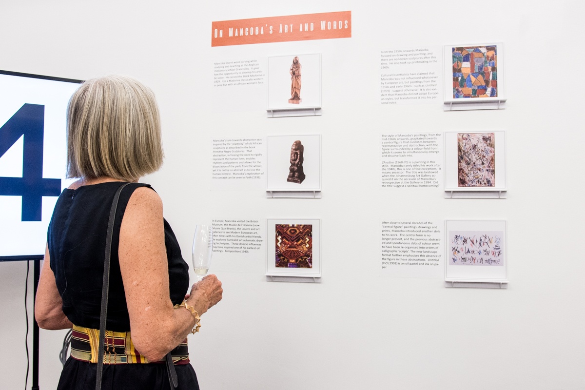 Event photograph from the Ernest Mancoba Symposium at A4 Arts Foundation. On the right, framed photographs of six of Mancoba's artworks are arranged on small shelves. On the left, a standing individual observes the arrangement on the wall.
