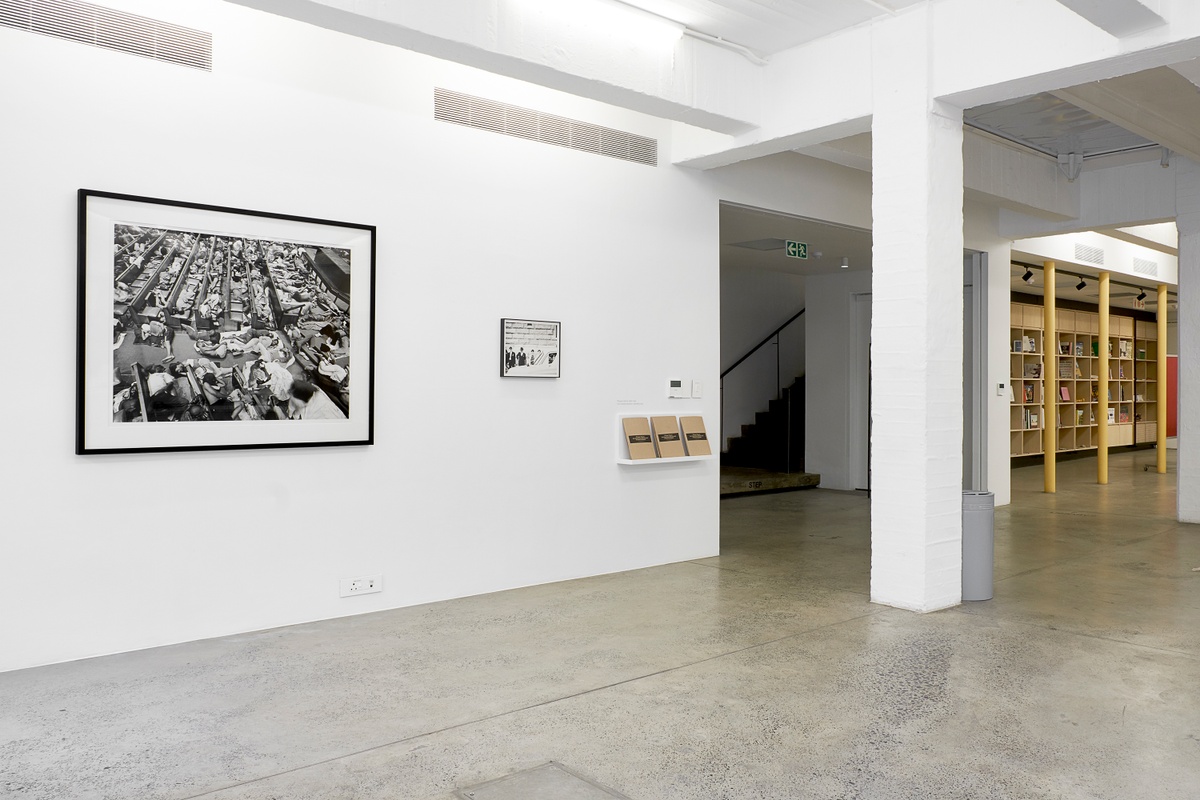 Installation photograph from the ‘Picture Theory’ exhibition in A4’s Gallery that depicts artworks in A4’s foyer. On the left, David Goldblatt’s monochrome photograph ‘Zimbabwe refugees given shelter in the Central Methodist Church, Johannesburg’ is mounted on the wall. In the middle, Goldblatt’s monochrome photograph ‘Policeman in a squad car on Church Square, Pretoria (Tshwane), Transvaal (Gauteng)’ in mounted on the wall, and a wall-mounted shelf next to it holds printed copies of the exhibition index.
