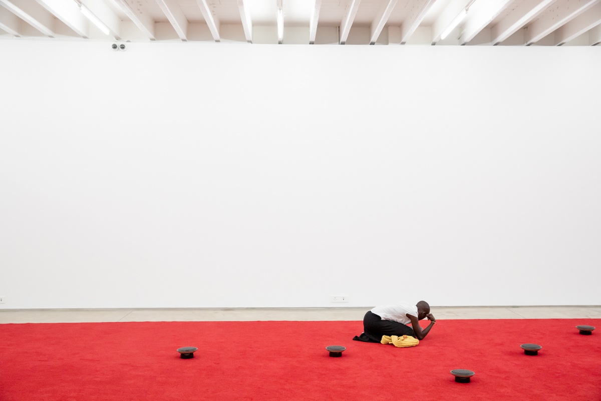 Event photograph from the opening of The Future Is Behind Us exhibition in A4’s gallery. At the front, James Webb’s sound installation ‘Prayer’ is visible, with red carpeting and upturned speakers. An individual is kneeling beside of the speakers to listen.
