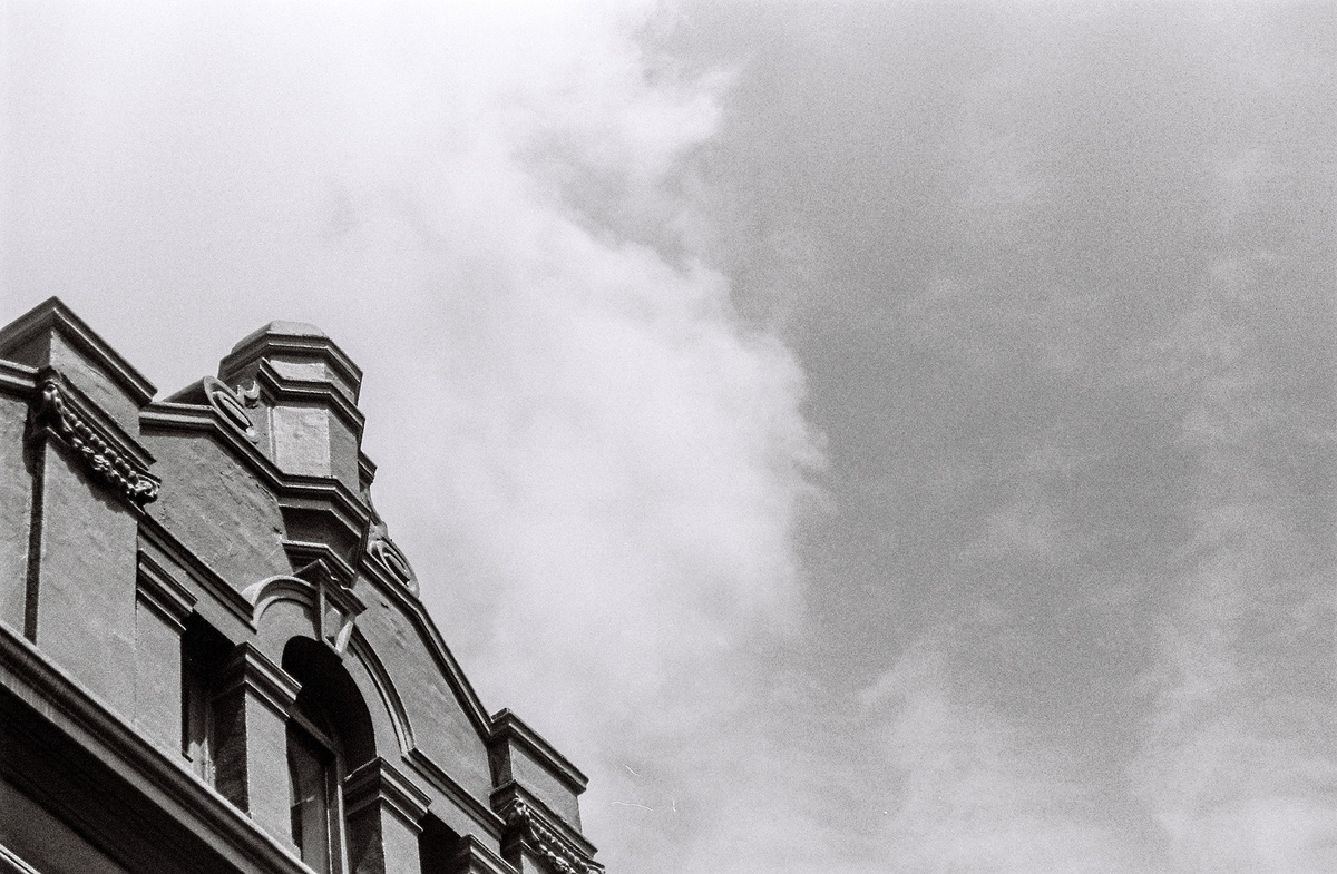 Monochrome event photograph from ‘Contact’, a collaboration between A4’s proto~ museum shop and Oath Magazine, taken by a participant. On the left, a detail of the top of a building. On the right, a view of a cloudy sky.
