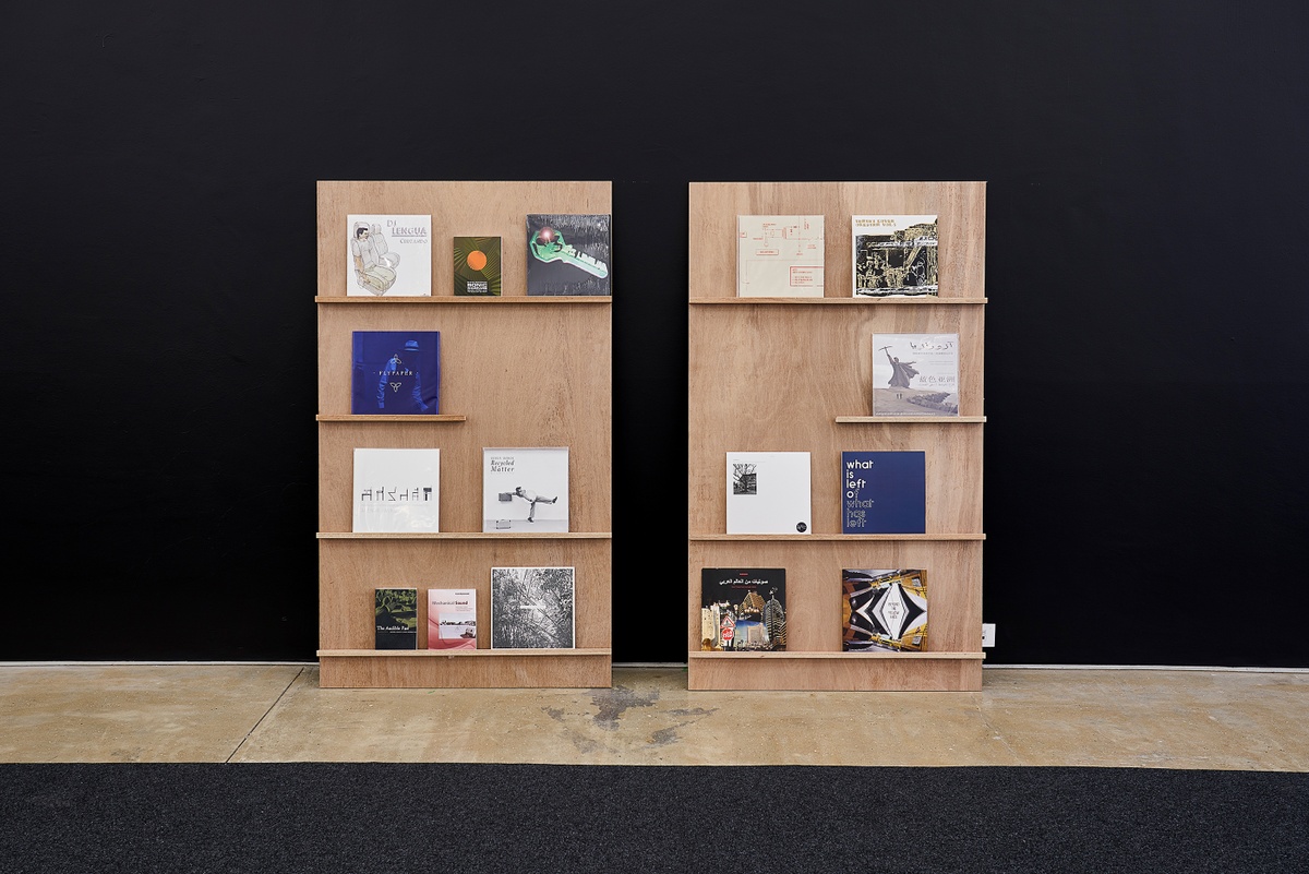 Installation photograph from the ‘Sounding the Void, Imaging the Orchestra V.1’ exhibition in A4’s Gallery. A selection of vinyl records from Bhavisha Panchia’s ‘Nothing to Commit Records’ collection and some printed matter is arranged on two rectangular wooden stands with narrow shelves.

