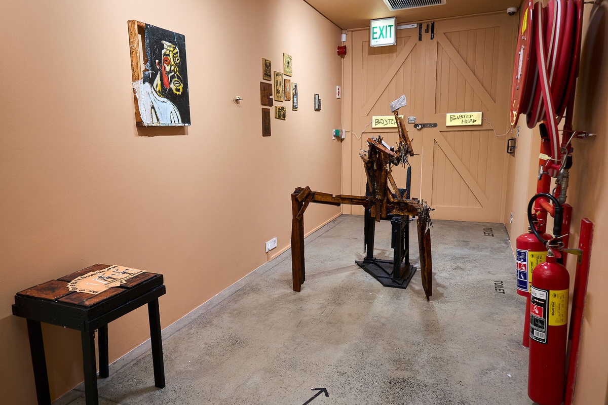 Installation photograph from Phillip Newman's exhibition in A4 Arts Foundation's Goods space. In the middle, a wood and metal sculpture sits on the concrete floor. On the left, a painting and drawings are mounted along the wall.
