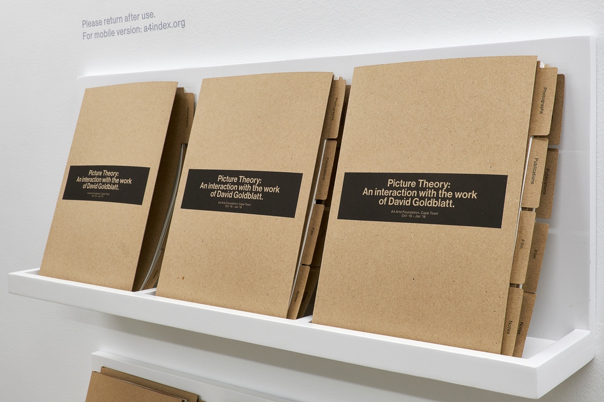 Photograph of the printed index for the ‘Picture Theory: An interaction with the work of David Goldblatt’ exhibition in A4’s Gallery. In the middle, brown paper folders with labeled tabs sit on a white shelf.
