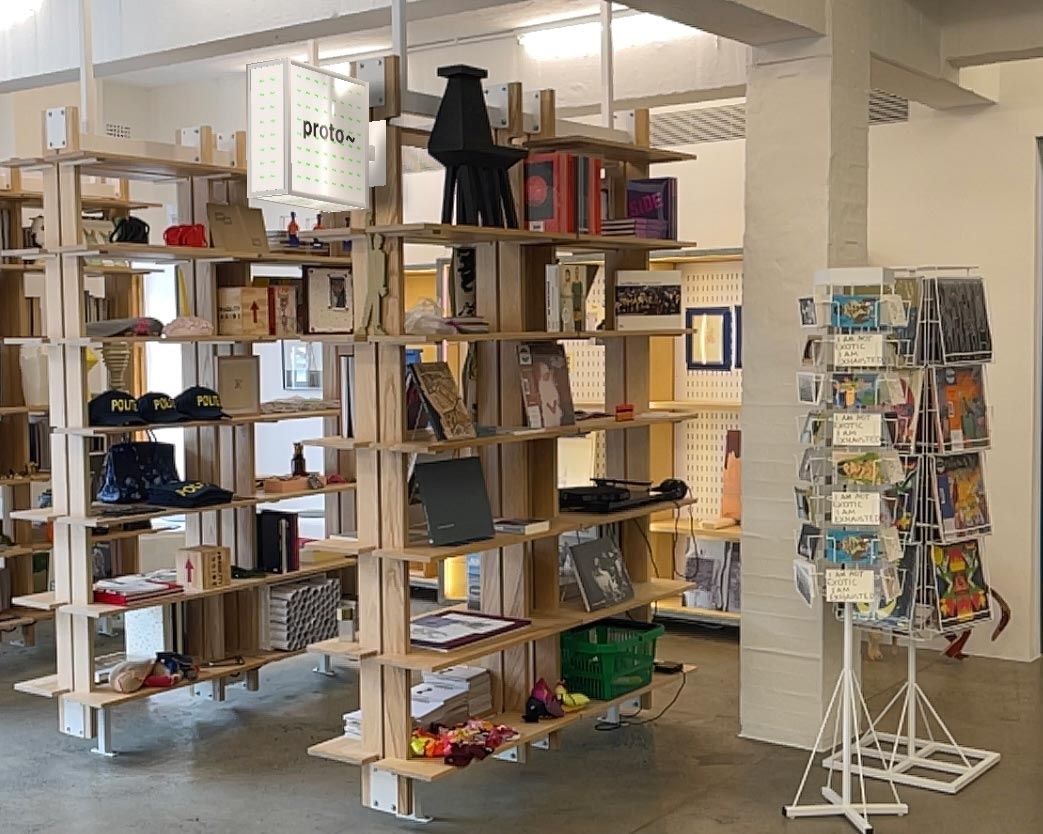 Install photograph from ‘Landscaping the Store,’ Meghan Ho-Tong’s redesign of A4’s Proto~ museum store. On the left, bookshelves feature books and small art objects. On the right, two rotating metal displays feature postcards and vinyl records. At the back, display cabinets with various art objects.
