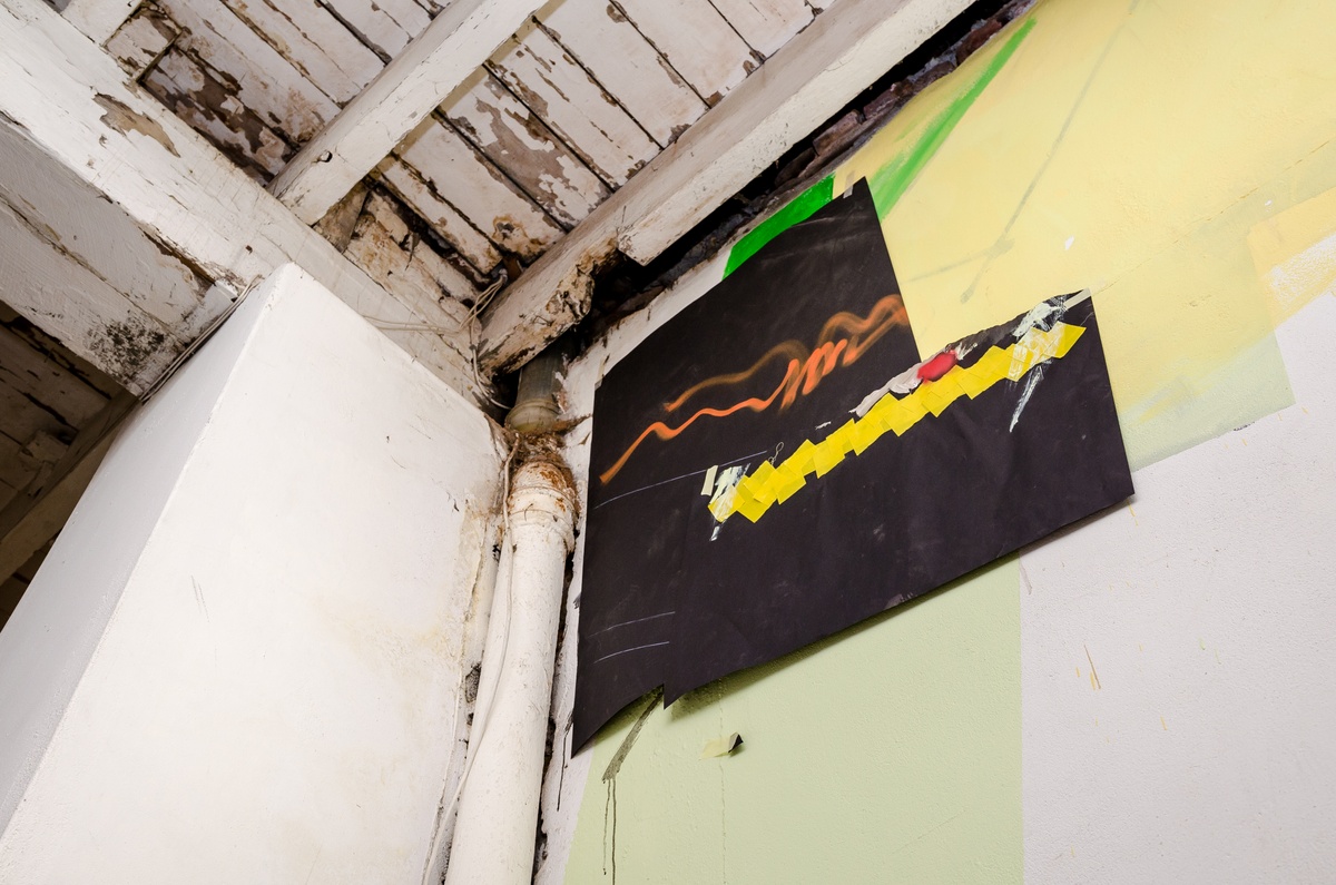 Process photograph from Dorothee Kreutzfeldt’s residency on A4’s 1st floor that depicts a corner where the wall meets the wooden ceiling, with piping and wiring from the floor above visible along the seem. On the right, a detail of a wall mural that consists of paint, drawing and irregular sheets of black material taped to the wall.
