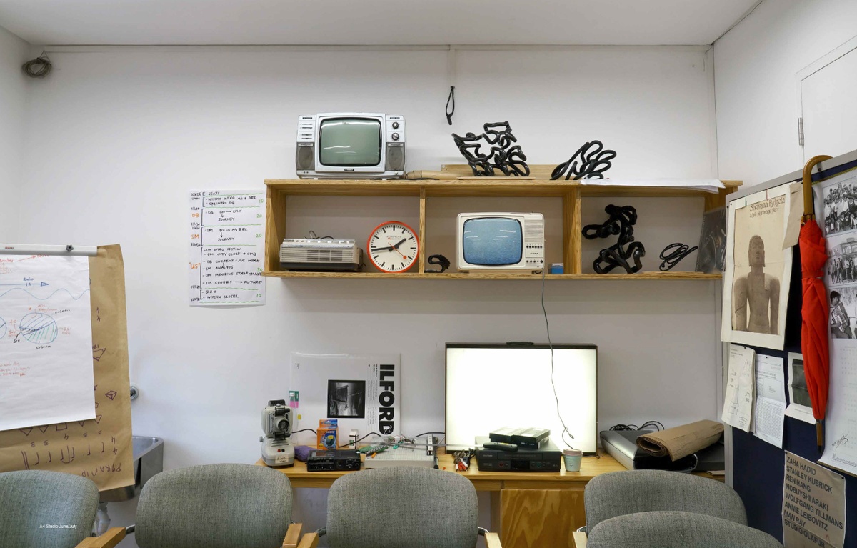 Process photograph from Sukuma Mkhize’s residency on A4’s top floor. At the back, the studio wall features wooden shelving and cupboards that host television sets, black sculptural objects and projectors. On the right and left, freestanding boards host notes and photographs.
