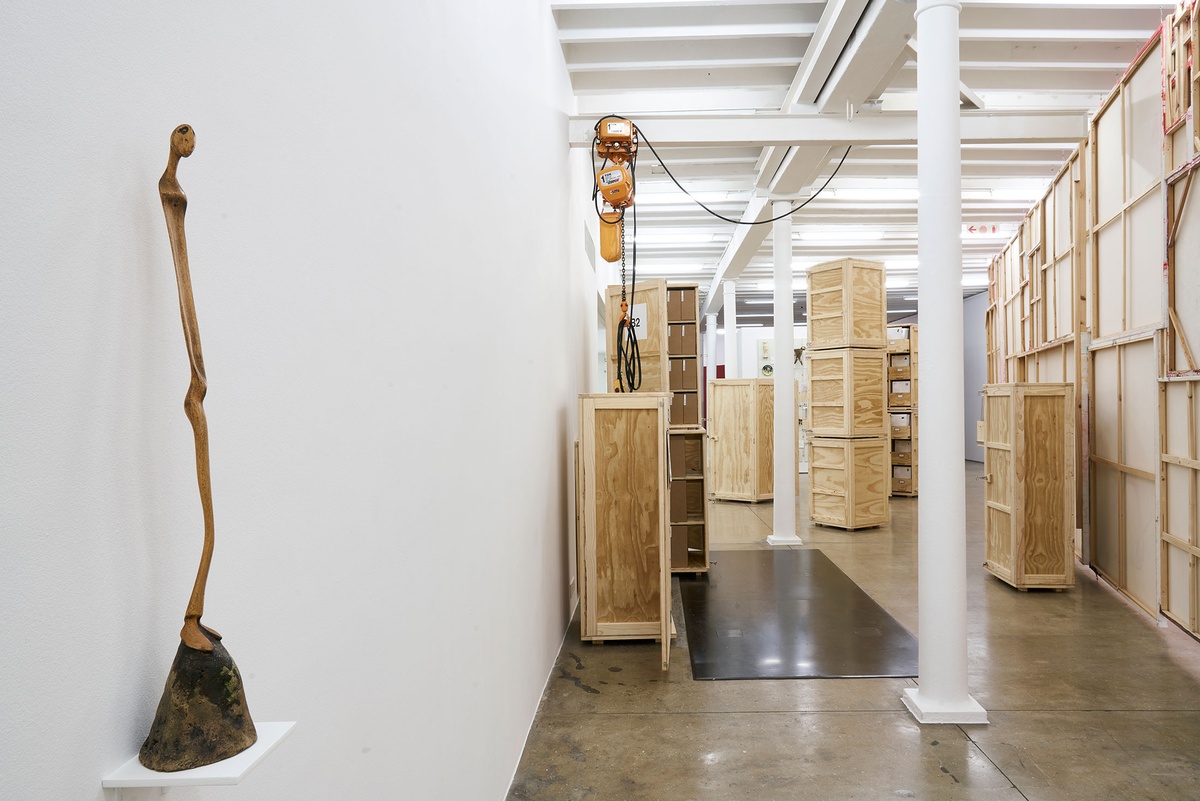 Installation photograph from the 'A Little After This' exhibition in A4 Arts Foundation's gallery. On the left, Lucas Sithole's carved yellowwood sculpture 'Not You!' sitting on a small wall-mounted shelf. On the right, collected objects from Penny Siopis' 'Will' work are stored in envelopes and wooden crates.
