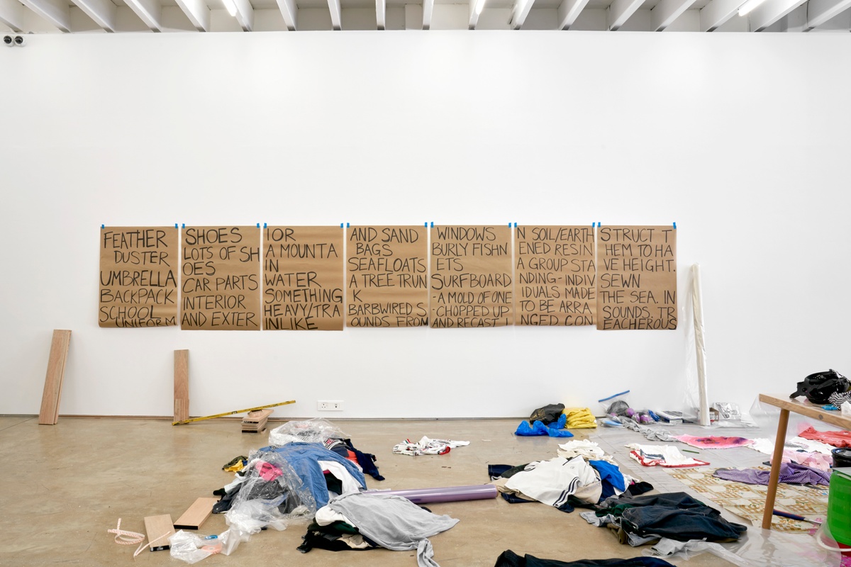 Process photograph from the ‘without a clear discernible image’ exhibition in A4’s Gallery, where the gallery was temporarily used as a studio space by the artist Kevin Beasley. At the back, a row of brown paper sheets is taped to the gallery wall, with writing that relates to the process and materials of Beasley’s practice.
