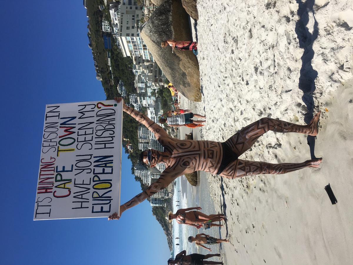 Event photograph from Kieron Jina’s ‘Catching Feelings, not Flights’ performance. The artist, their partially nude body covered in geometric designs, walks next to a Cape Town beach carrying a canvas that reads ‘It’s hunting season in Cape Town, have you seen my European husband?’ 
