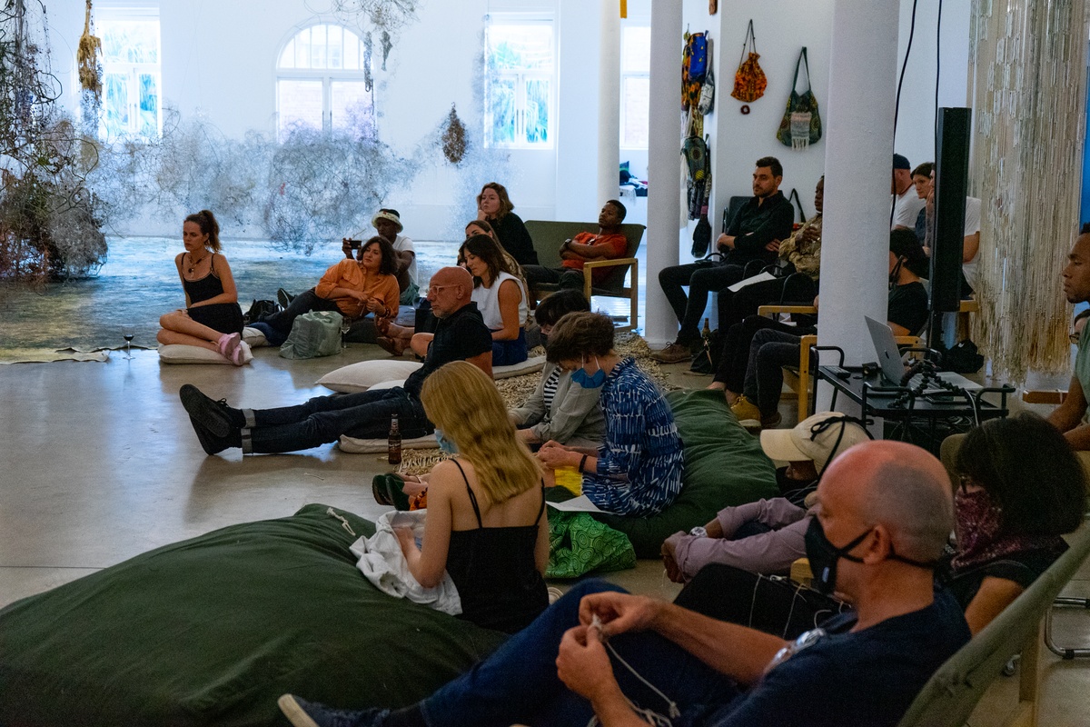 Event photograph from ‘Knots’, a conversation between Igshaan Adams and Josh Ginsburg, marking the close of ‘Open Production’, Adams’ hybrid studio/exhibition in A4’s Gallery. On the right, attendees are seated on cushions, chairs and couches.
