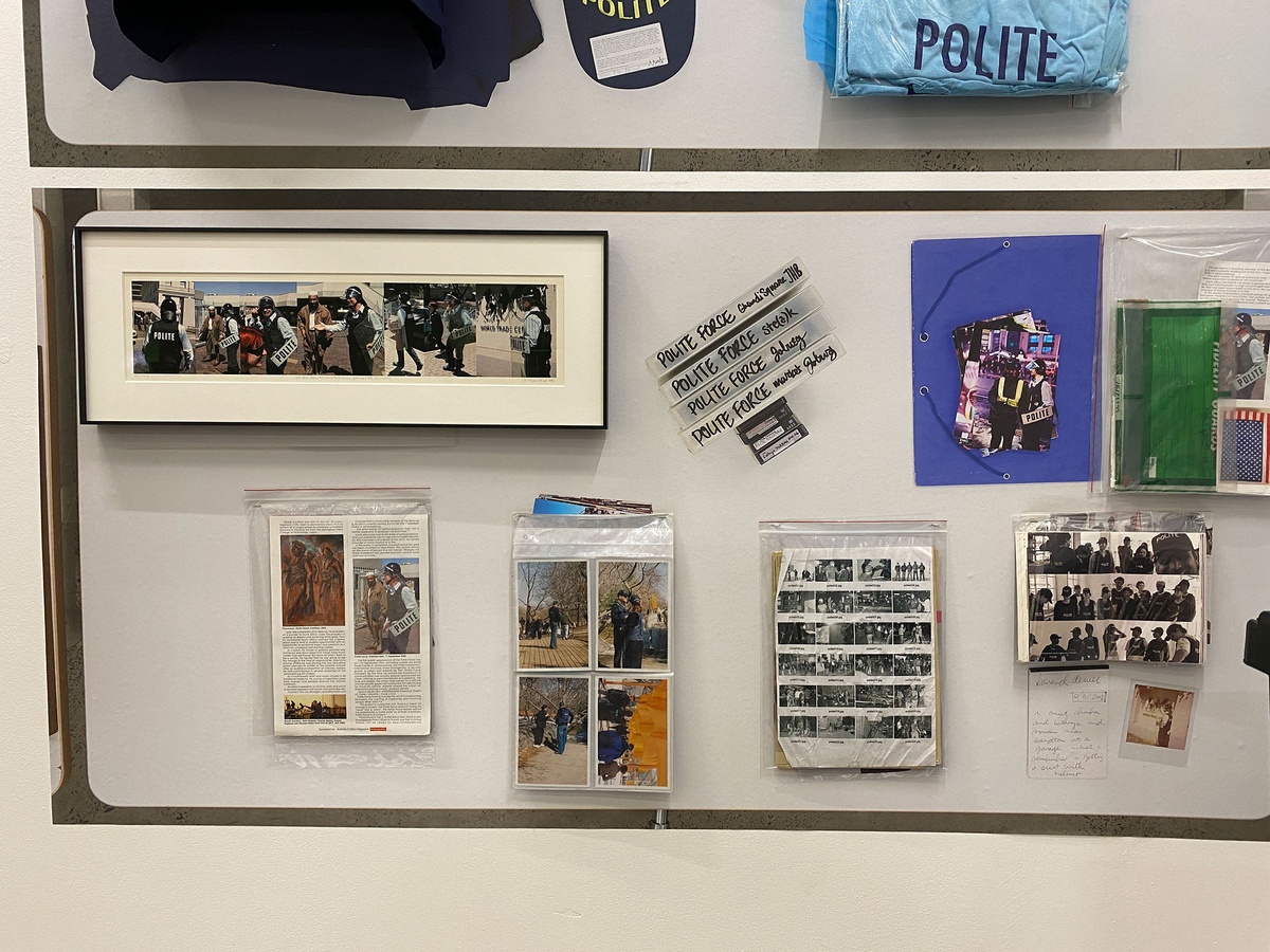 Event photograph from A4’s booth at the 2022 Investec Cape Town Art Fair. A large rectangular photographic print on the wall shows a topdown view of the unpacking of Christian Nerf’s ‘Polite Force’ archive. Objects from the archive are mounted on the wall so as to align with their photographic traces.
