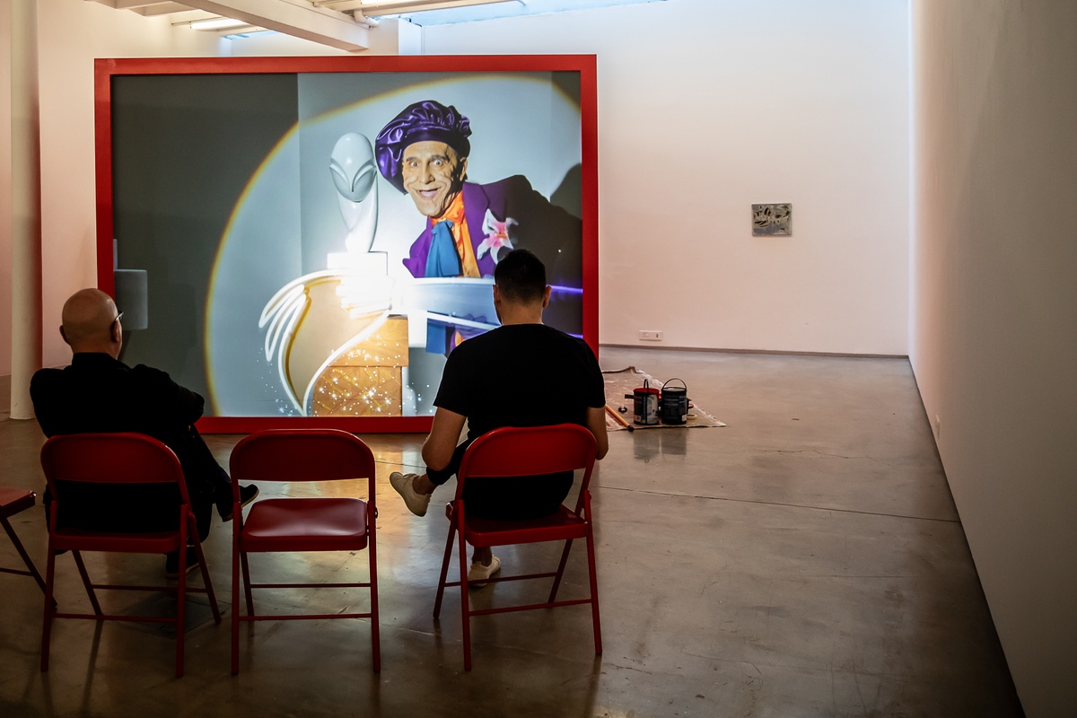 Event photograph from the preview of the 'A Little After This' exhibition in A4 Arts Foundation that shows Alex Da Corte's video installation 'ROY G BIV'. At the back, a large red wooden box with a back-projected screen plays Da Corte's video. At the front, 7 red powder-coated viewing chairs are arranged in an arch, with attendees seated there.
