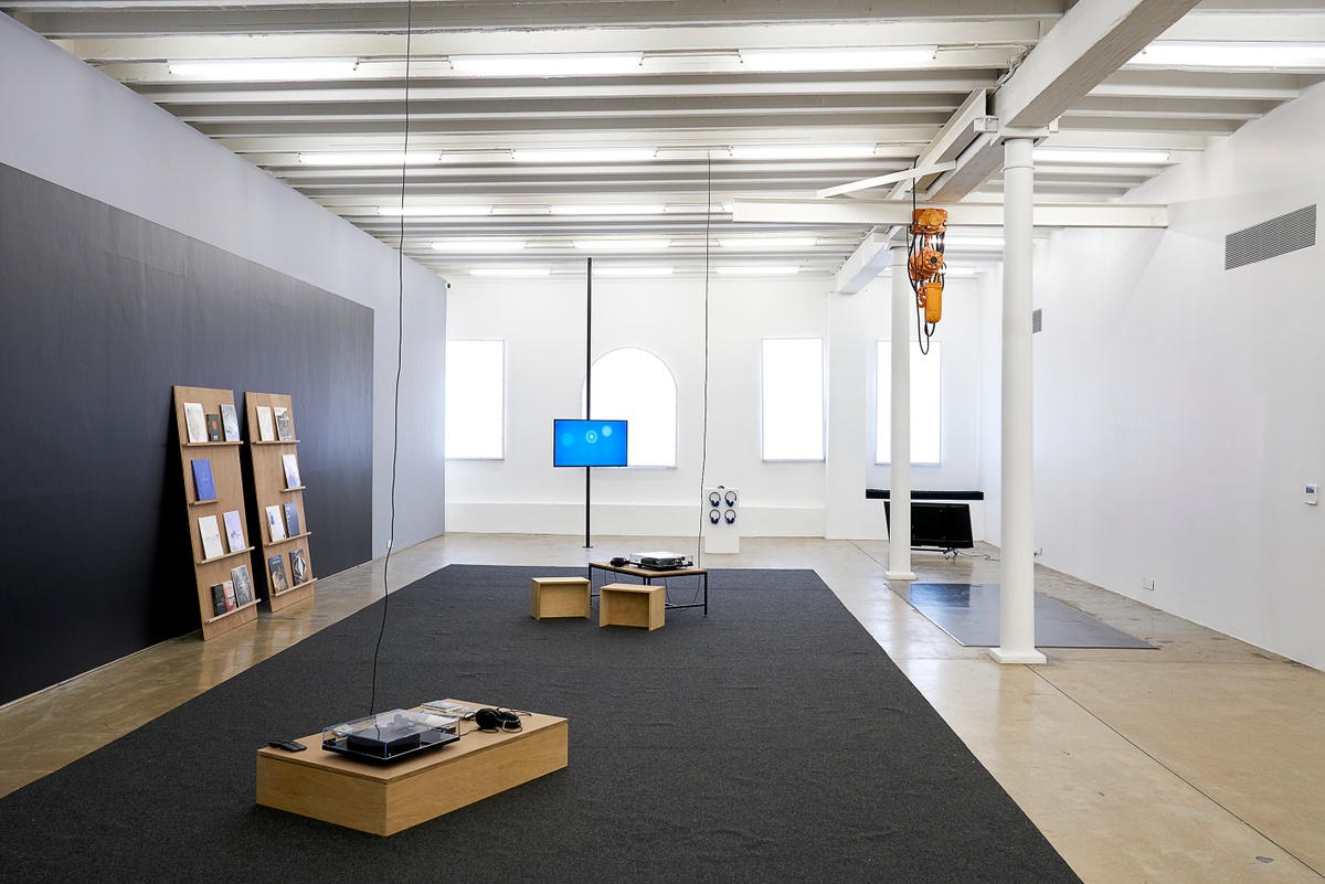 Installation photograph from the ‘Sounding the Void, Imaging the Orchestra V.1’ exhibition in A4’s Gallery. In the middle, two listening stations consist of low wooden tables with record players and headphones. On the left, a selection from Bhavisha Panchia’s collection of vinyls and books ‘Nothing to Commit  Records’ is arranged on two wooden shelving units. At the back, Jenna Sutela & Ville Haimala’s web-based installation ‘eAR’ and Sutela’s video ‘Talking Machine’ is displayed on screen on the floor.
