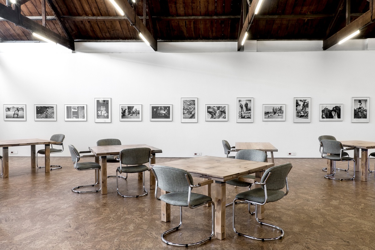 Installation photograph from the launch of Omar Badsha’s book ‘Seedtimes’ on A4’s top floor. At the front, wooden tables with seating is arranged along the room. At the back, a row of framed monochrome photographs line the white wall.
