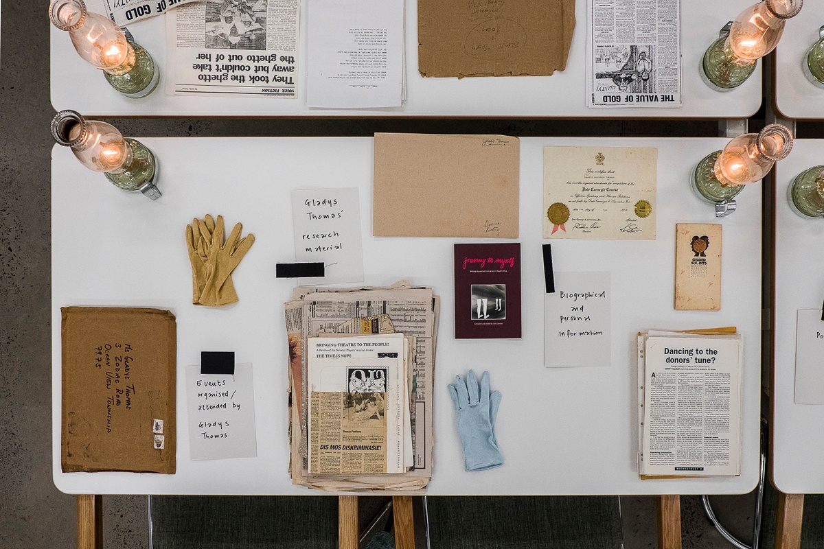 Installation photograph from the ‘Gladiolus’ exhibition on A4’s ground floor. The topdown view shows a selection of printed matter arranged on a white table along with gloves and lit glass lamps.
