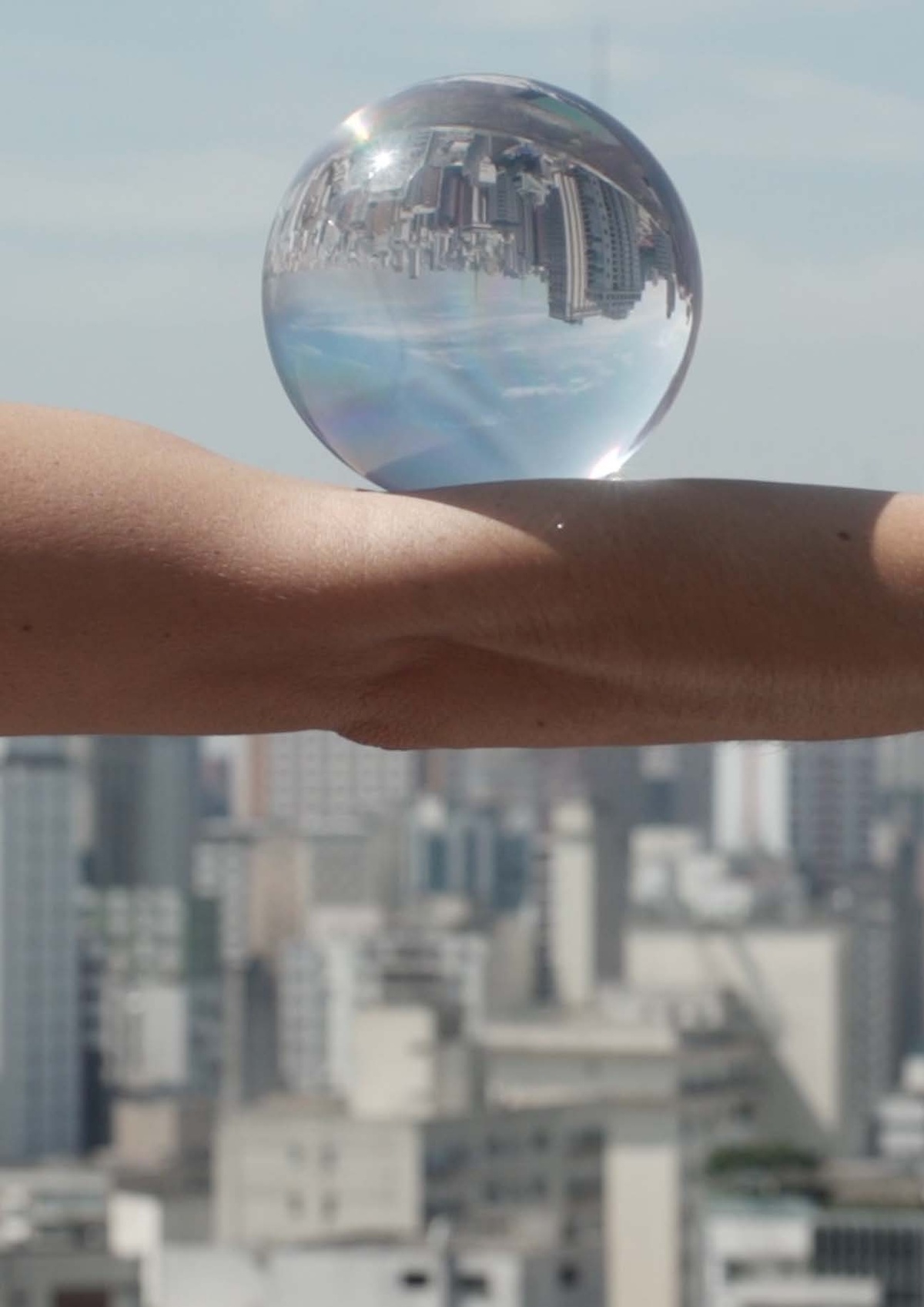 Process image from the ‘How to Remain Silent’ exhibition on A4’s ground floor that consists of a still frame from Lia Chaia’s video performance ‘Aleph’. At the front, a closeup view of an arm with a glass orb balancing on it. At the back, an out of focus view of São Paulo cityscape.
