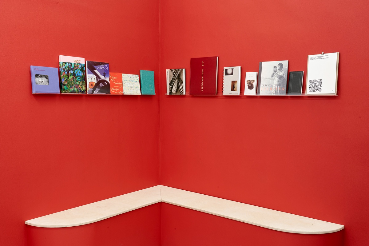 Installation photograph of the You to Me, Me to You exhibition that shows a corner of the gallery with the walls painted red. At the top, a selection of books are arranged on narrow perspex shelves. At the bottom, wooden benches are attached to the base of the wall.
