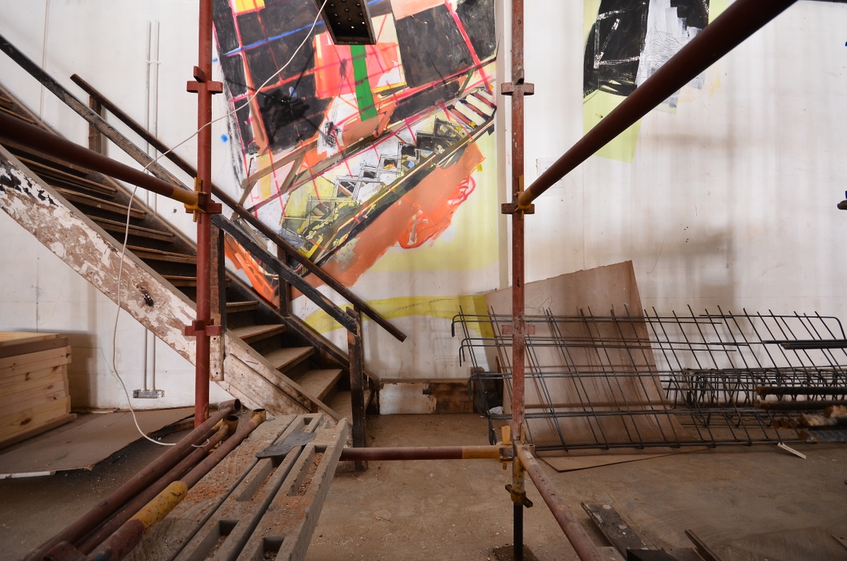Process photograph from Dorothee Kreutzfeldt’s residency on A4’s 1st floor. At the back, a painted mural on a white wall, with a a wooden staircase in front of it. At the front, metal scaffolding holds concrete beams.
