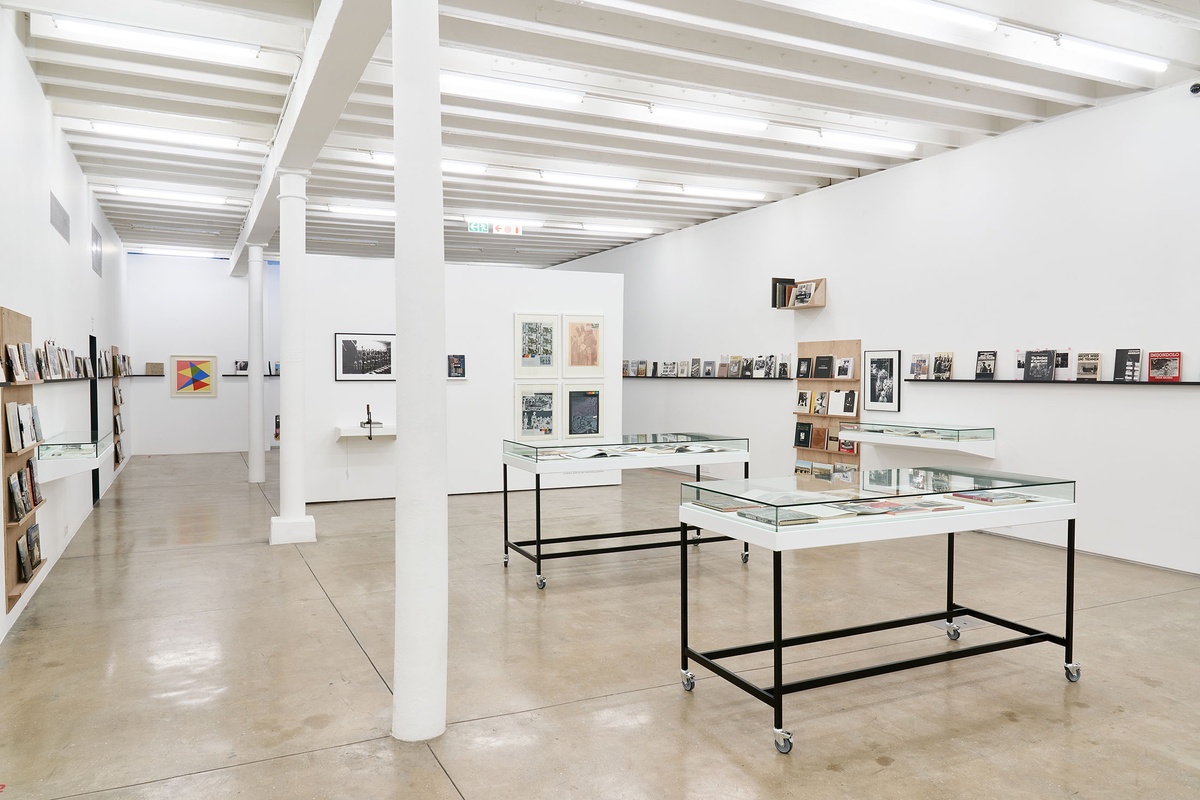 Installation photograph from the 'Photo Book! Photo-Book! Photobook!’ exhibition in A4’s Gallery. At the front, two freestanding display cases with printed matter. In the middle, a moveable gallery wall that features Peter Magubane’s photograph ‘Mine recruits humiliated by being forced to strip naked for tuberculosis examination, Witwatersrand (1968)’, Jacobsens Publishers’ book ‘Index of Objectionable Literature’, Ernest Cole’s book ‘House of Bondage’ and four screenprints from Gavin Jantjes’ ‘A South African Colouring Book’ series. On the left, shelving units with printed matter from the years 1945 to 1967 line a white wall, with Albert Newall’s oil painting ‘Harmonic Development within a Square’ mounted on a wall at the back. On the right, shelving units with printed matter line a white wall, along with David Goldblatt’s photograph ‘Climbing into kibbles at the shaft-head, the shift gets ready to go down. President Steyn No.4 shaft, Welkom, Orange Free State. 1969’.
