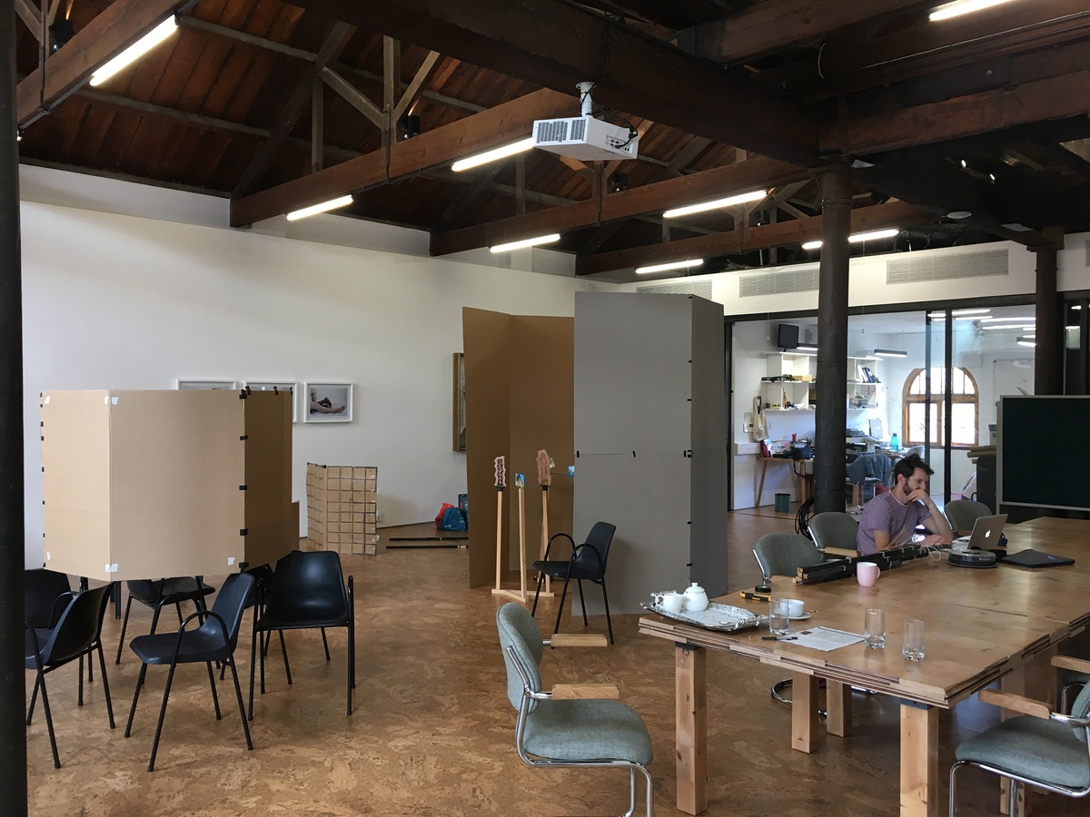 Process photograph from Jonah Sack’s residency on A4’s top floor. On the right, Sack sits working on a laptop at a wooden table. On the left, cardboard constructions of various sizes are arranged throughout the space.
