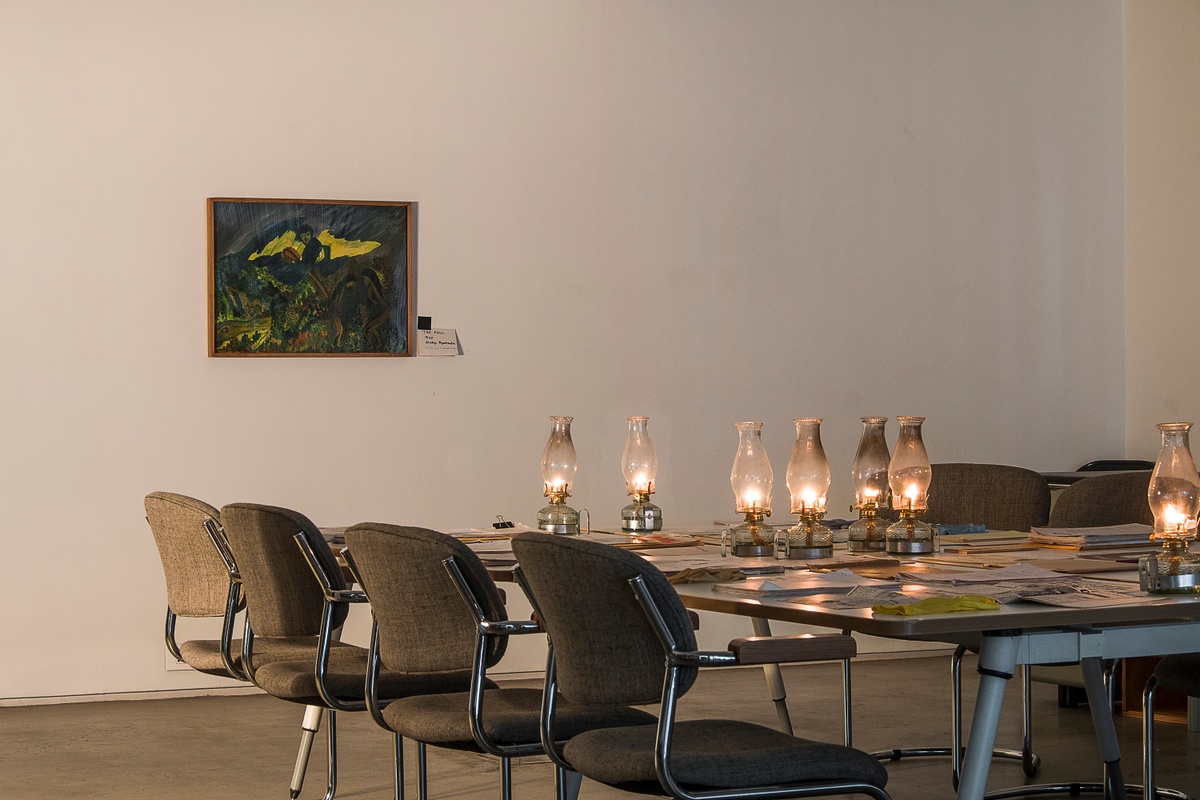 Installation photograph from the ‘Gladiolus’ exhibition on A4’s ground floor. At the back, Gladys Mgulandlu’s gauche painting ‘The Fall’ is mounted on a white wall. At the front, tables with printed matter and oil lamps.
