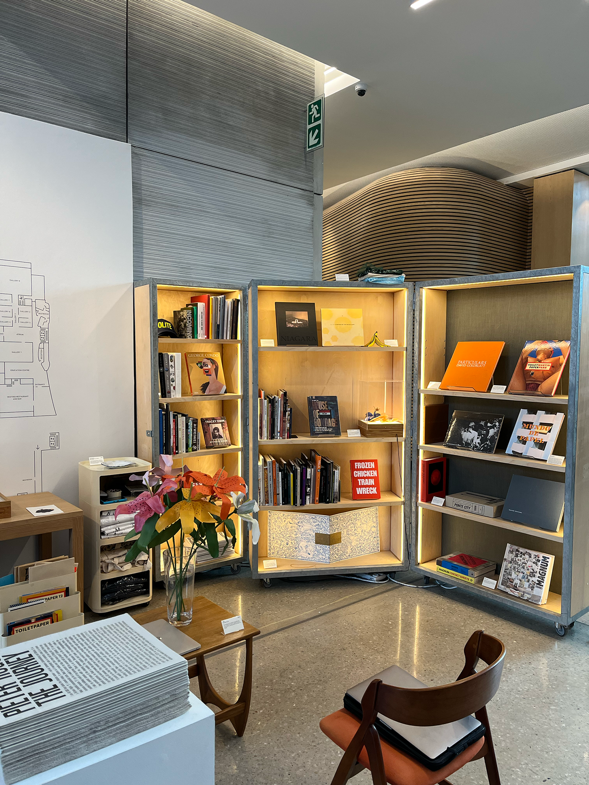 Installation photograph of Proto's booth at the 2022 Cape Town Art Book Fair. At the back, a wood and metal display cabinet holds a selection of books. At the front, a white plinth features a stack of Pieter Hugo's newsprint publication 'The Journey'.
