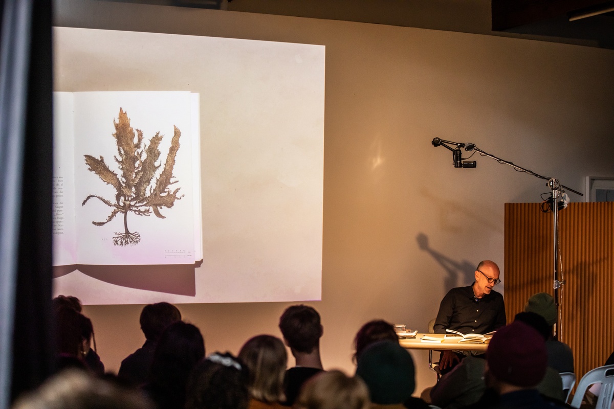 Event photograph from the book launch of 'ArtThrob: 25 Years of Art Writing in South Africa' in A4 Arts Foundation. On the right, writer Sean O'Toole is seated at the speaker's table. On the left, a livestream top-down view of the speaker's table is projected onto the wall.
