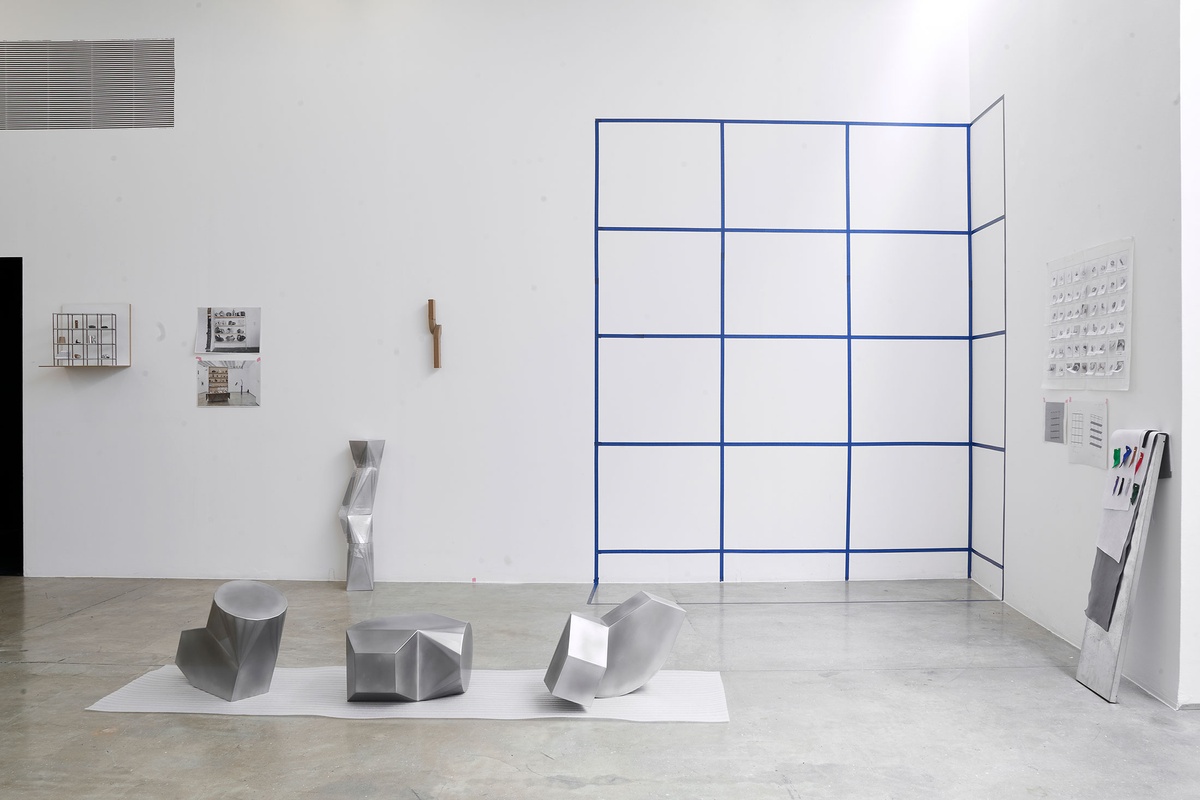 Process photograph of Kyle Morland’s working space during the 2nd rendition of ‘Parallel Play’ in A4’s Gallery. At the front, three metallic sculptural forms sit on plastic foam sheeting. At the back, the layout for a shelf is mapped out in the corner of the gallery with blue tape.
