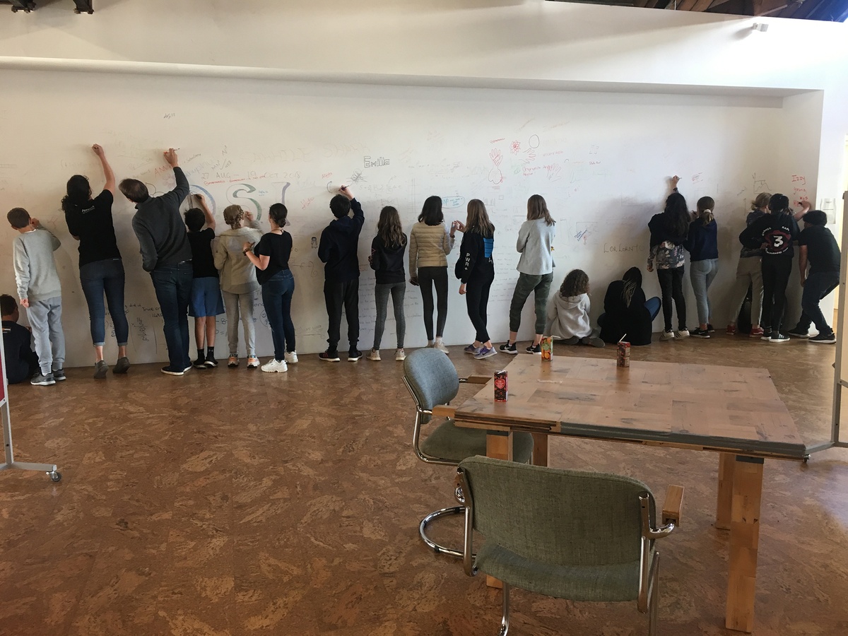 Photograph from the ‘Water Workshop with Edu Africa’ exchange in A4’s Gallery. At the front, a square wooden table with juice boxes on A4’s top floor. At the back, a row of students drawing and writing on a white wall.

