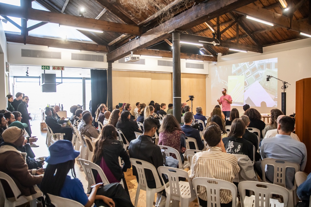 Event photograph from the book launch of 'ArtThrob: 25 Years of Art Writing in South Africa' in A4 Arts Foundation. At the front, attendees are seated in rows. At the back, Josh Ginsburg addresses the attendees.
