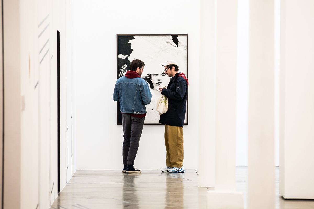 Event photograph from opening of the Customs exhibition in A4’s Gallery that shows two attendees standing in front of Dor Guez’s framed photographic print ‘Samira’ mounted on a white gallery wall.
