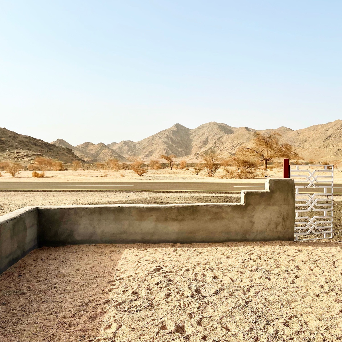 Process photograph from Waiting Upon, Sumayya Vally’s Course of Enquiry at A4, depicting a roadside mosque in Saudi Arabia. At the front, a low-wall enclosure with a gate. In the middle, a tarred road. In the back, various mountains.
