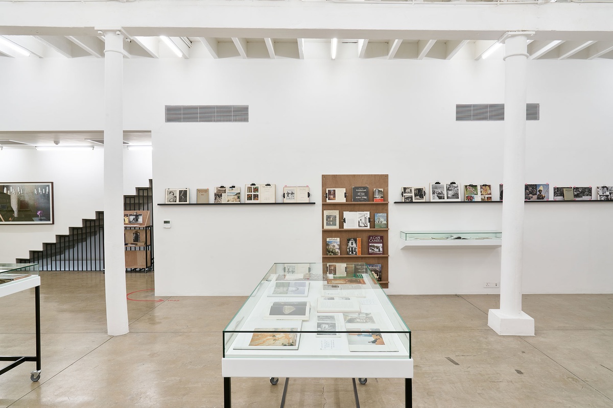 Installation photograph from the Photo Book! Photo-Book! Photobook! exhibition in A4’s Gallery. At the front, a freestanding glass display case with printed matter. At the back, shelving units with printed matter from the years 1945 to 1967 line a white gallery wall.
