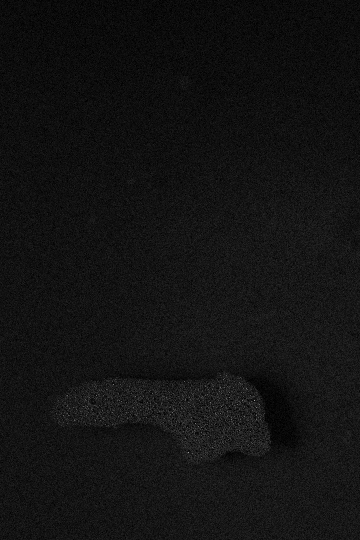 A monochrome photographic print from Thembinkosi Hlatshwayo’s series ‘wanders about’ that depicts liquid with bubbles on a black surface.
