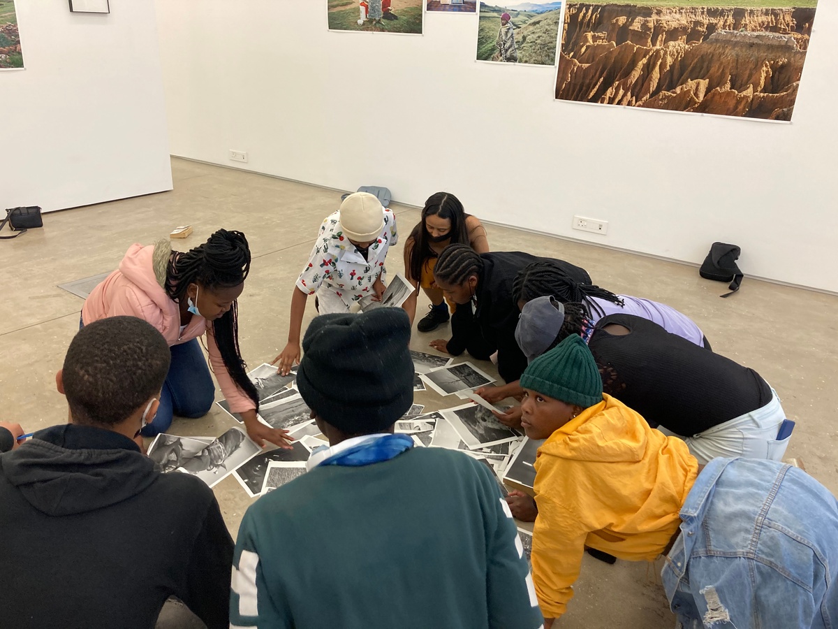 Event photograph from the ‘Lalela x A4' exchange during the ‘Tell It to the Mountains’ exhibition in A4’s Gallery. Lalela students and facilitators are huddled around monochrome photocopies of Lindokuhle Sobekwa’s photographs on the gallery floor.
