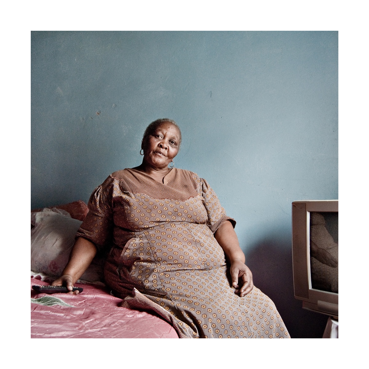 Photograph from Jabulani Dhlamini’s residency on A4’s top floor shows an individual seated on the edge of a bed on the left, with a television screen to the right.
