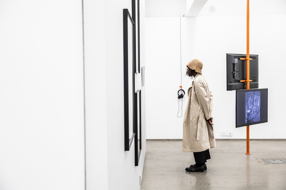 Event photograph from the opening of the ‘Model’ exhibition in A4’s Reading Room. An attendee stands with their back to a support beam that hosts two counter facing video screens, with Tony Yanick’s video ‘BOBBI LYNN’ visible on one of them.
