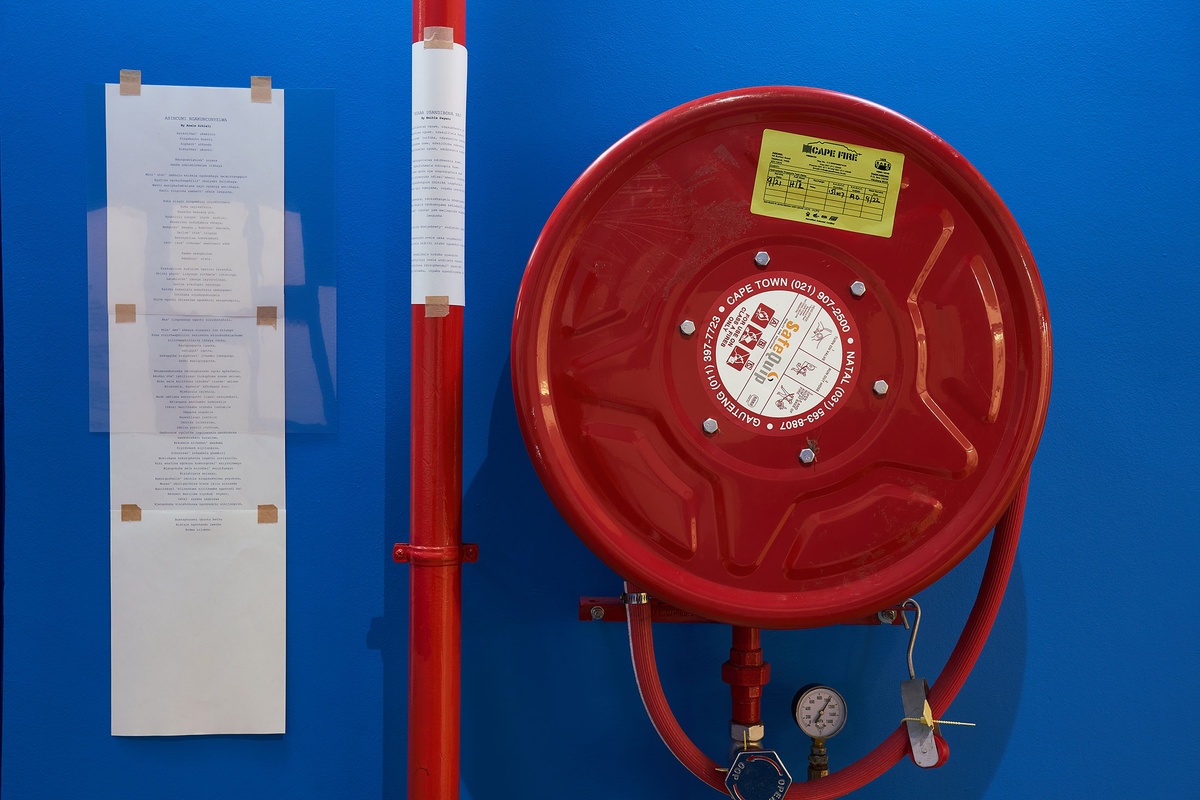 Installation photograph from the ‘Umcimbi’ exchange in A4’s Goods project space. On the left, printed poems are pasted onto the wall next to a fire hose.
