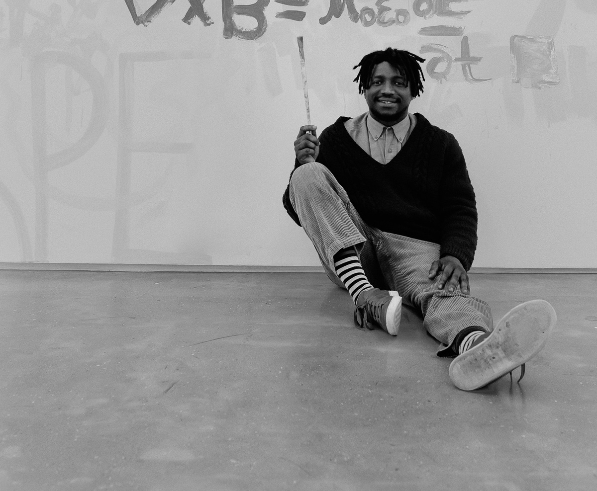 Ephemera from Sukuma Mkhize’s residency on A4’s top floor. At the front, a monochrome photograph shows Mkhize seated at the base of a wall holding a paint brush. At the back, the wall features painted formula.

