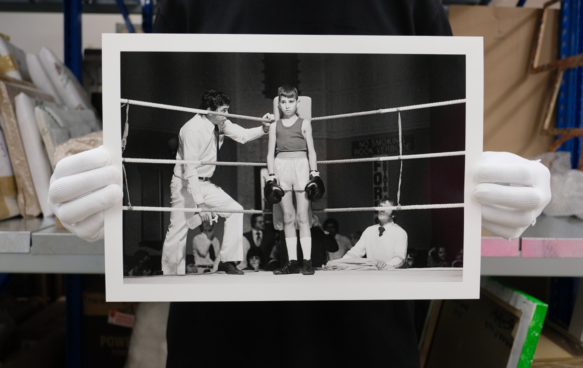 Process photograph from the ‘Picture Theory’ exhibition in A4’s Gallery. Hands wearing archive gloves hold a printed copy of David Goldblatt’s photograph ‘Before the fight: amateur boxing at the Town Hall, Boksburg’. In the middle, a young boy stands in a the corner of a boxing ring. On the left, a man stands in attendance outside the ring.
