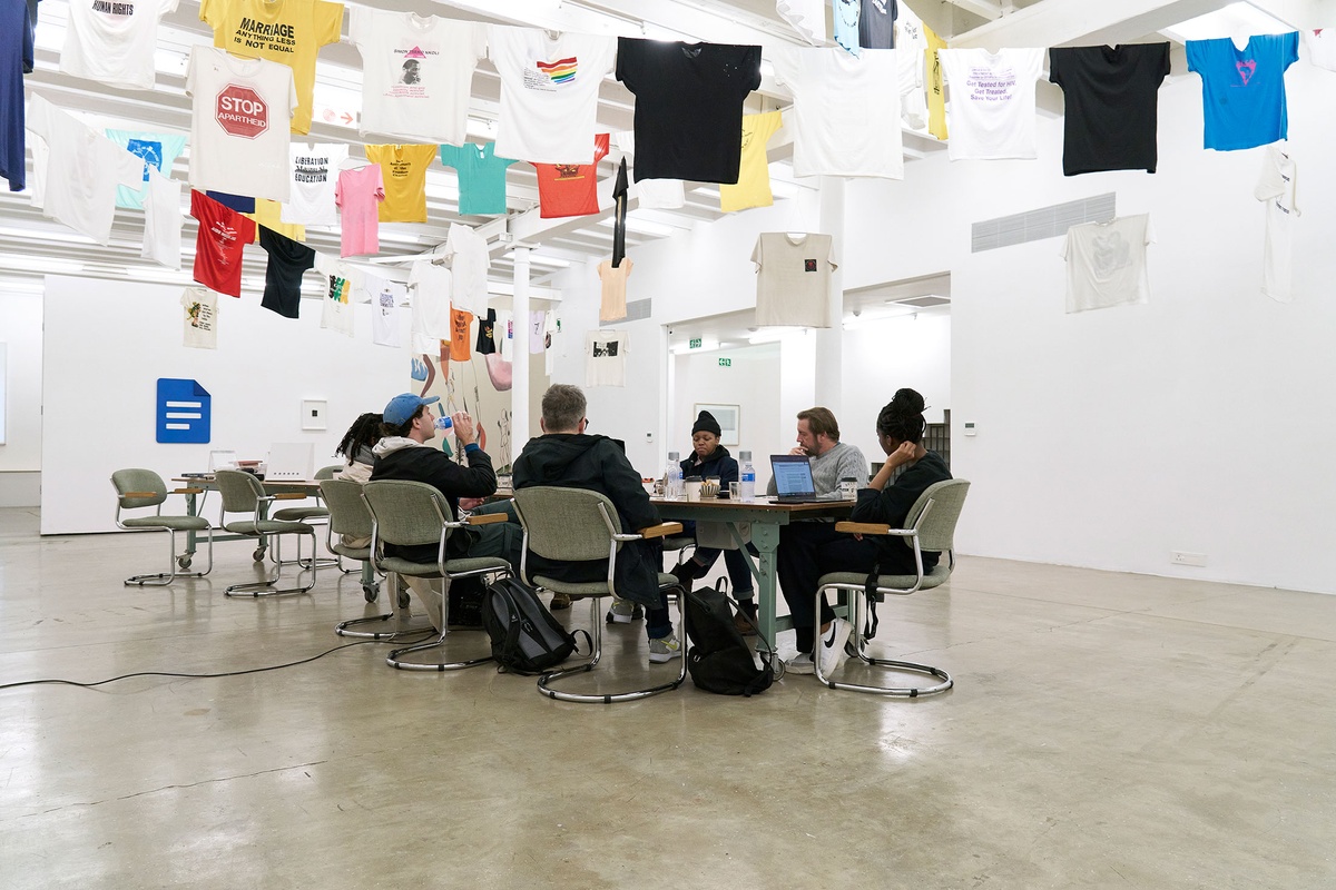 Event photograph of the roundtable discussion for the Common exhibition in A4 Arts Foundation's Gallery. In the middle, roundtable participants are seated around a long wooden table. Above, t-shirts from the GALA Queer archive and South African History Archive are suspended on criss-crossing lines.
