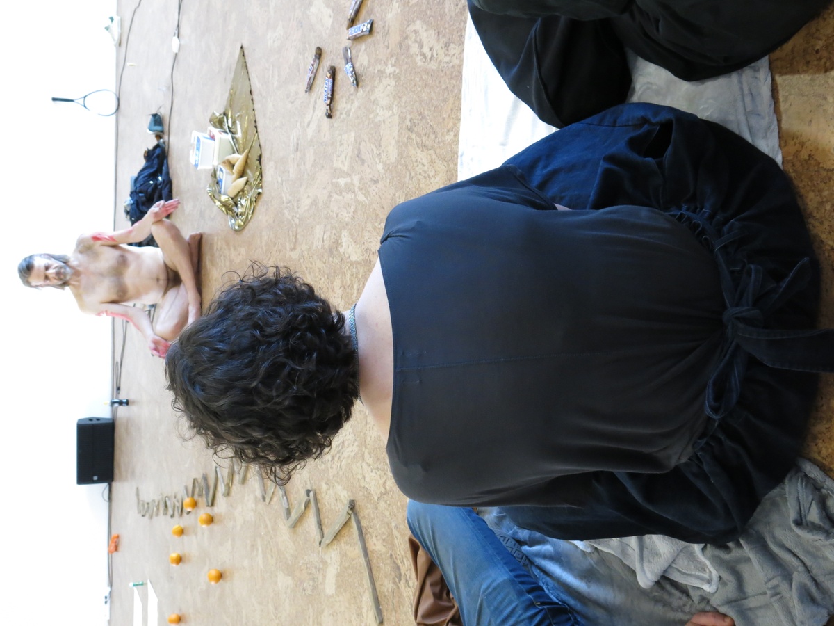 Event photograph from Mårten Spångberg’s solo dance ‘Digital Technology’ on A4’s top floor. At the back, Spångberg is sitting unclothed and crosslegged on the floor among various objects. At the front, an attendee is seated on the floor.
