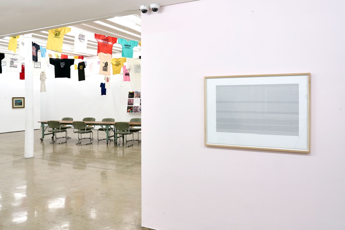 Installation photograph of the Common exhibition. Above on the left, T-shirts with slogans from the GALA Queer Archive and SAHA are suspended on criss-crossing lines. In the middle at the back are framed Fabian Saptouw prints from their ‘ISBN University’ series with layered ISBN and ISSN numbers, hung on a pale pink wall.
